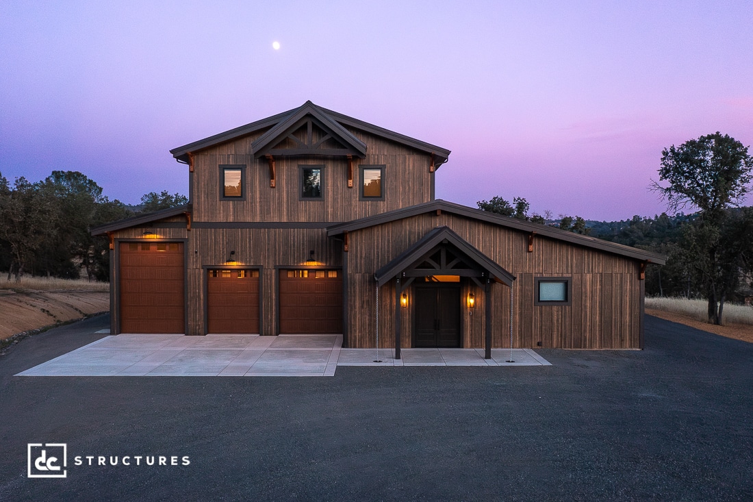 A modern barn-style house with wooden siding, three garage doors, and warm exterior lights, sits under a purple dusk sky with a visible moon. Trees and grass surround the stone driveway.