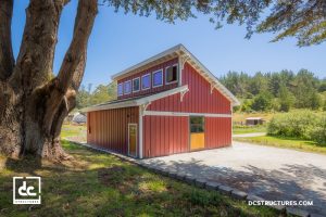 A modern red barn with white trim sits on a concrete pad surrounded by green grass and trees under a clear blue sky. The barn has large windows and a wooden door, with forested hills in the background.