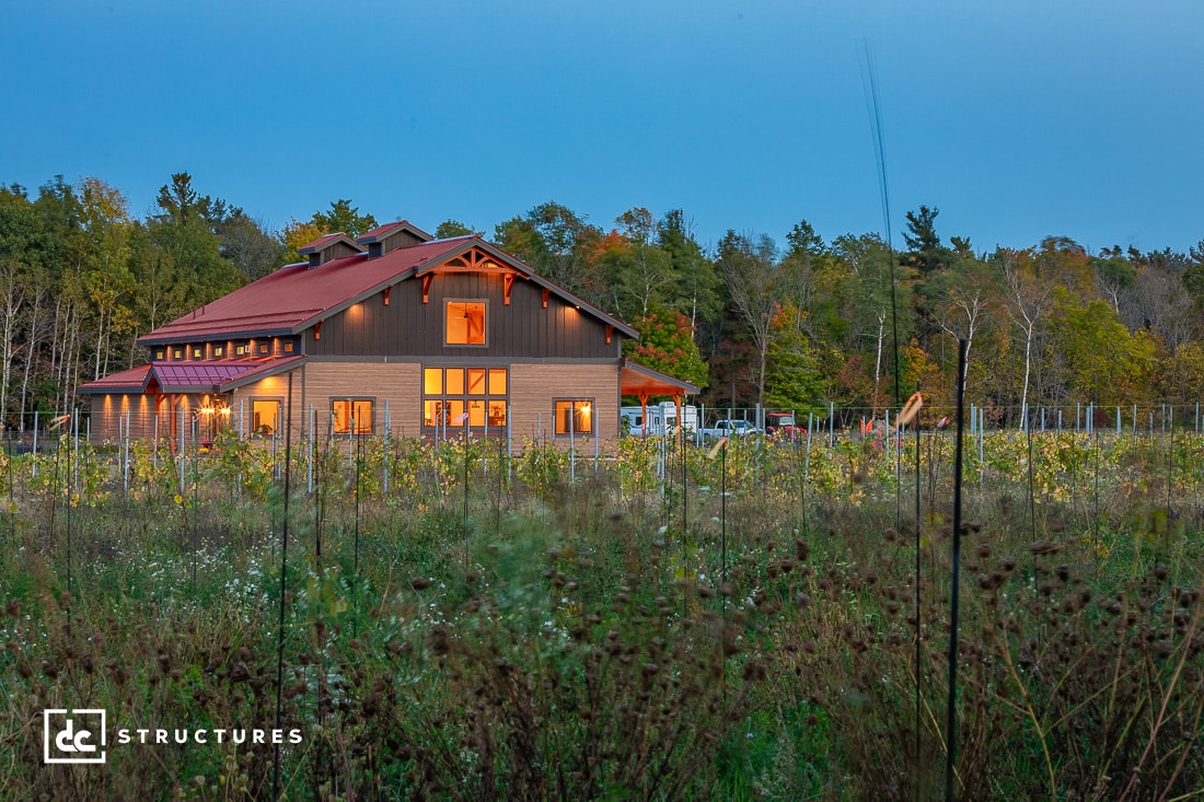 A large, modern barn-style building with warm lights is surrounded by a field of wildflowers and trees at dusk. The building features wood and dark trim accents, with a clear sky in the background.