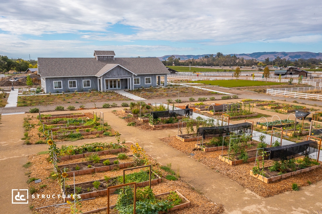 A large community garden with raised beds filled with plants sits in front of a gray barn-style building under a partly cloudy sky, surrounded by fields, trees, and white fences.
