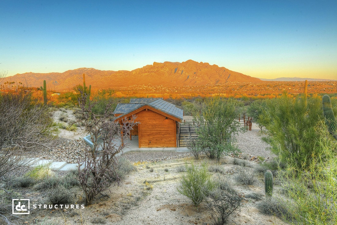 A small wooden cabin sits among desert plants and cacti, with a vast mountain range in the background under a clear blue sky. The scene is bathed in warm, golden sunlight.