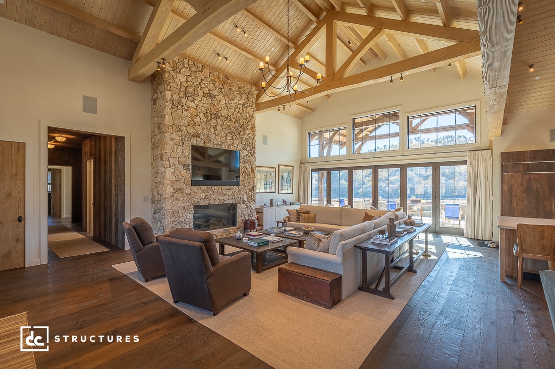 Spacious living room with exposed beam ceiling, stone fireplace, cozy sofas, big windows, wood floors, rustic decor, outdoor view.