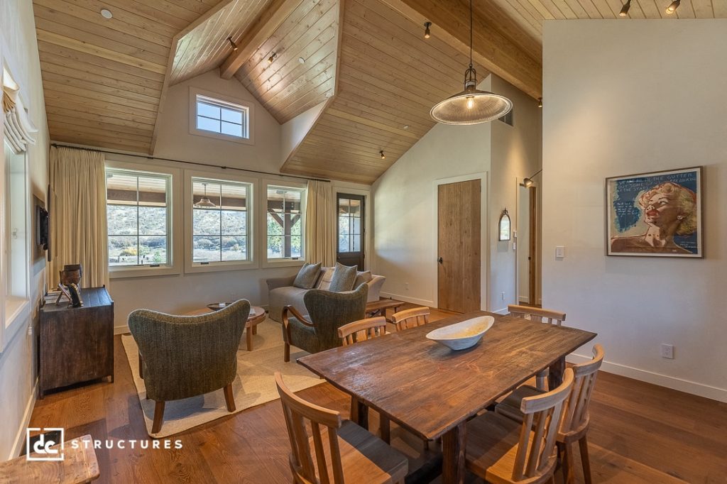 A bright, rustic living and dining area with wood ceilings, large windows, a wooden table and chairs, cozy armchairs, and a wall portrait.