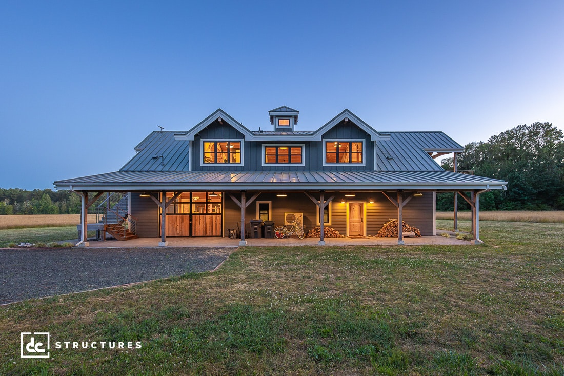 A large, two-story modern barn home with a metal roof, wide wraparound porch, and warmly lit windows set in a grassy field at dusk.