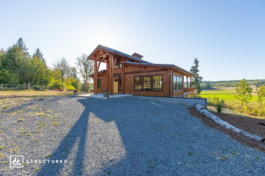 A modern wooden house with large windows sits on a gravel driveway, surrounded by green grass, trees, and open landscape under a clear blue sky.
