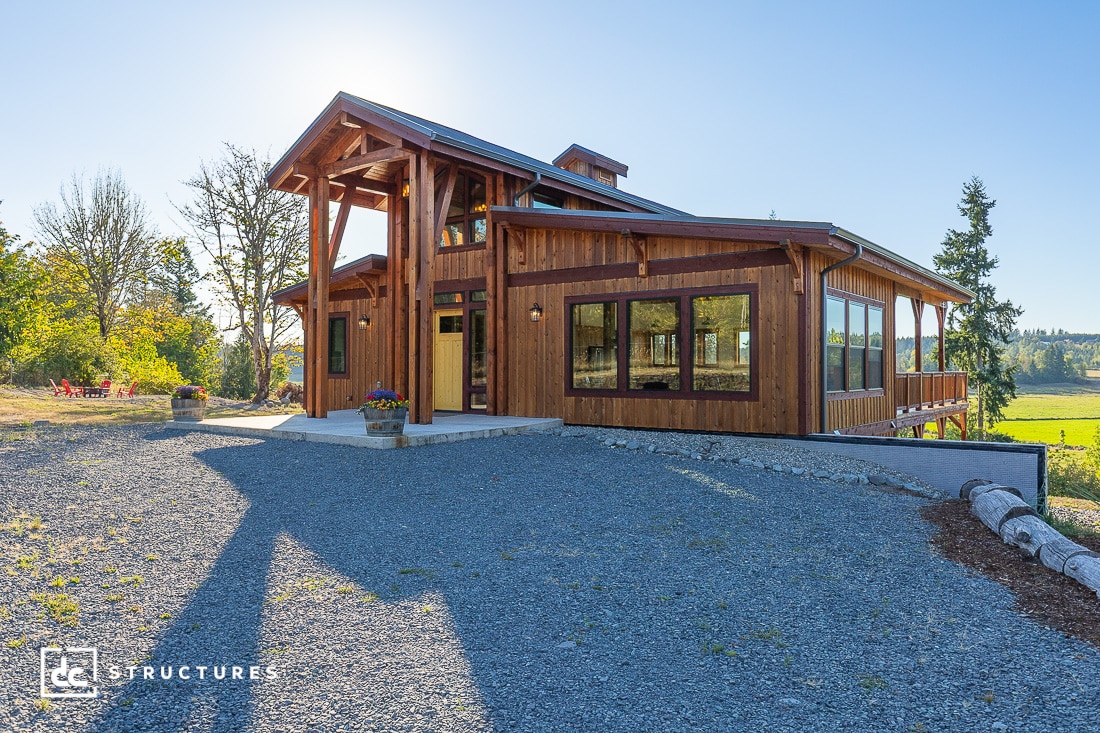 A modern wooden house with large windows and a covered porch on a gravel driveway, surrounded by green fields and trees.