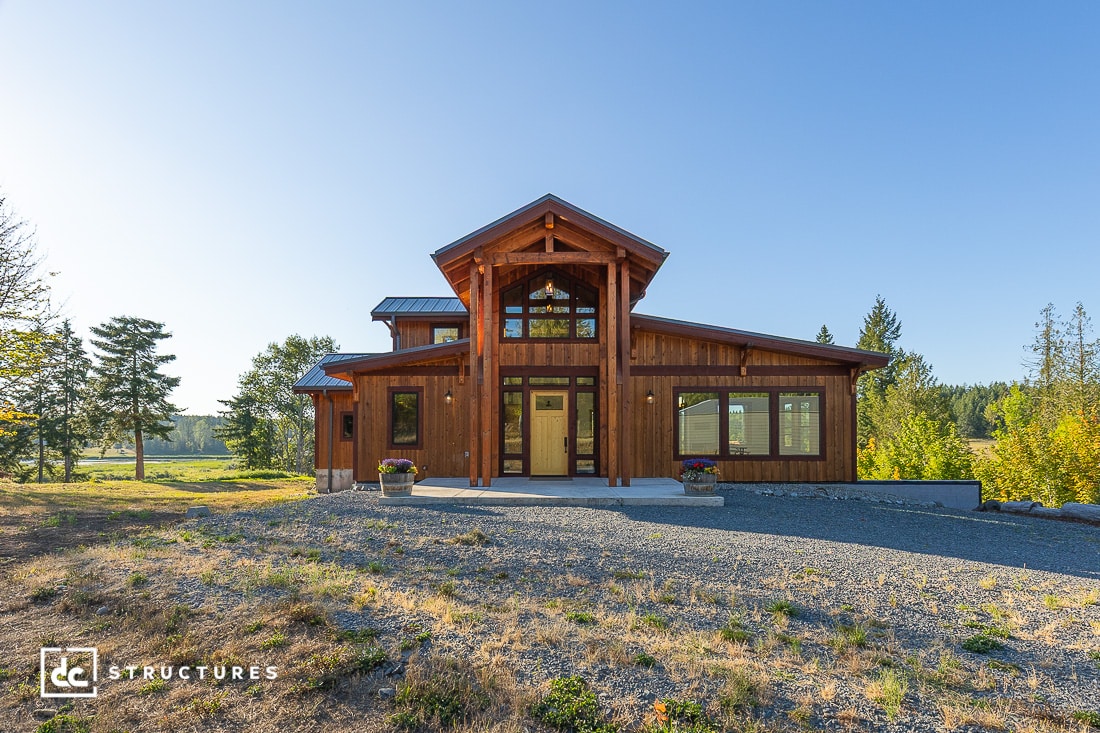 A wooden barn-style house with large windows and a covered entryway sits on a gravel driveway amid grass, trees, and blue sky.