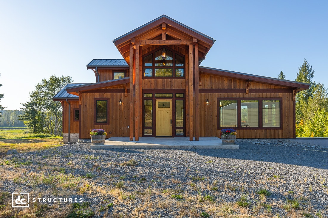 A modern wooden house with large windows and a peaked roof, surrounded by greenery, sits on a gravel driveway under a clear sky. Two potted plants flank the entrance.