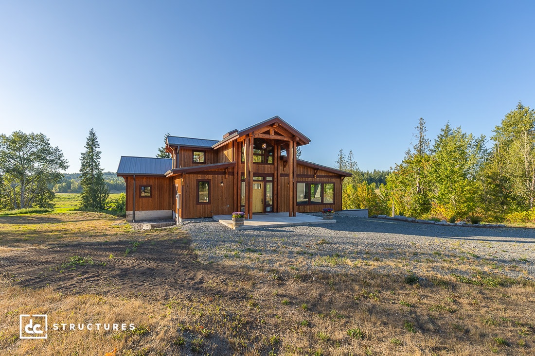 A modern wooden house with large windows and a covered porch on a gravel driveway, surrounded by trees and grass under a blue sky.