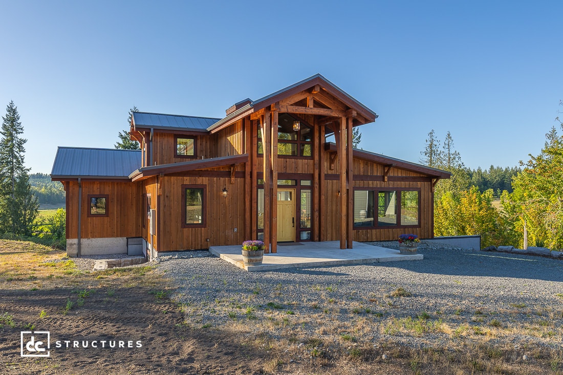 A modern wooden house with large windows and a covered front porch in a rural area, featuring a metal roof and gravel driveway.