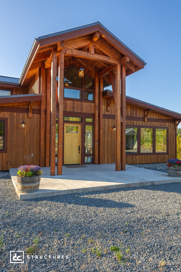 A modern wooden house with a tall covered entrance, large windows, and a gravel driveway. Two flower pots flank the front door.