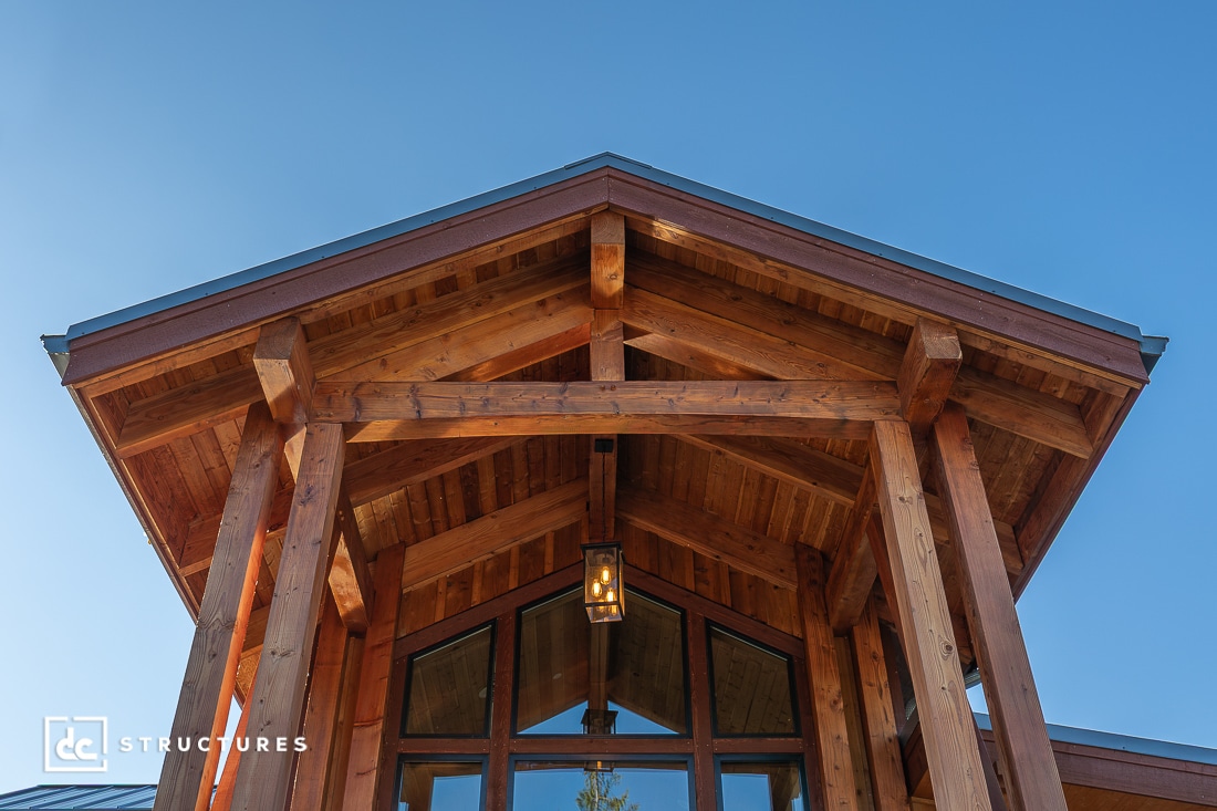 A close-up, low-angle view of a timber frame structure with exposed wooden beams, a peaked roof, and a hanging lantern against a clear blue sky.