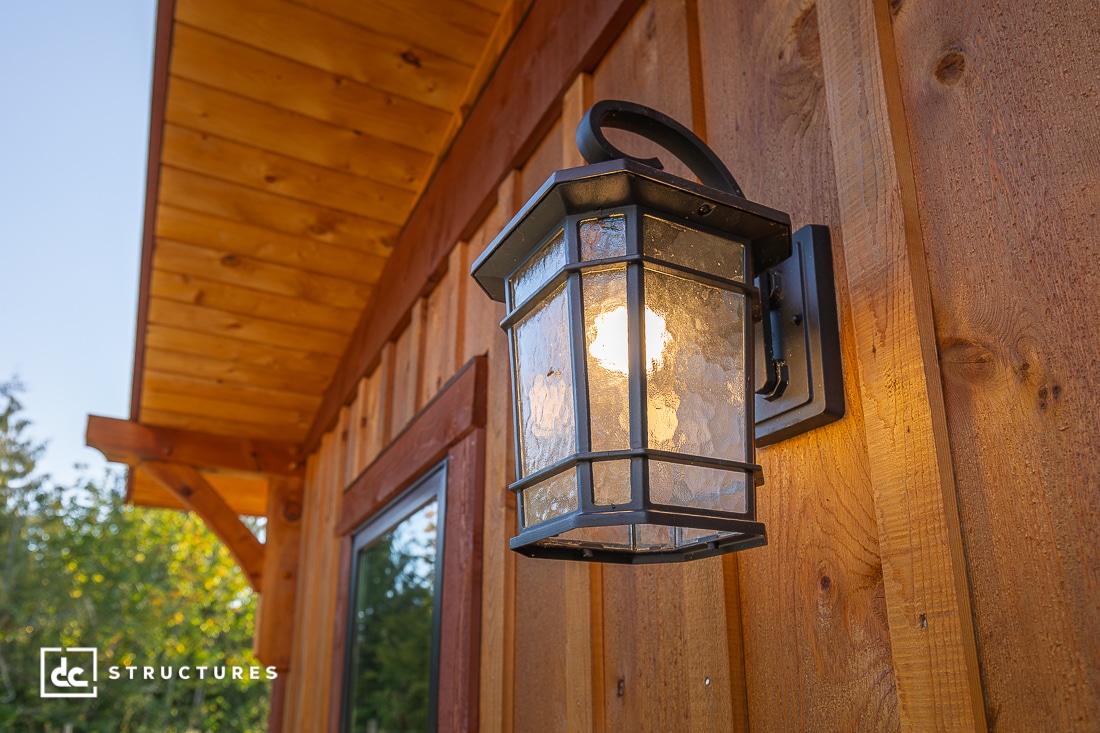 A black metal outdoor lantern is mounted on a wooden wall, illuminated in sunlight. A wooden overhang, window, and trees are visible.
