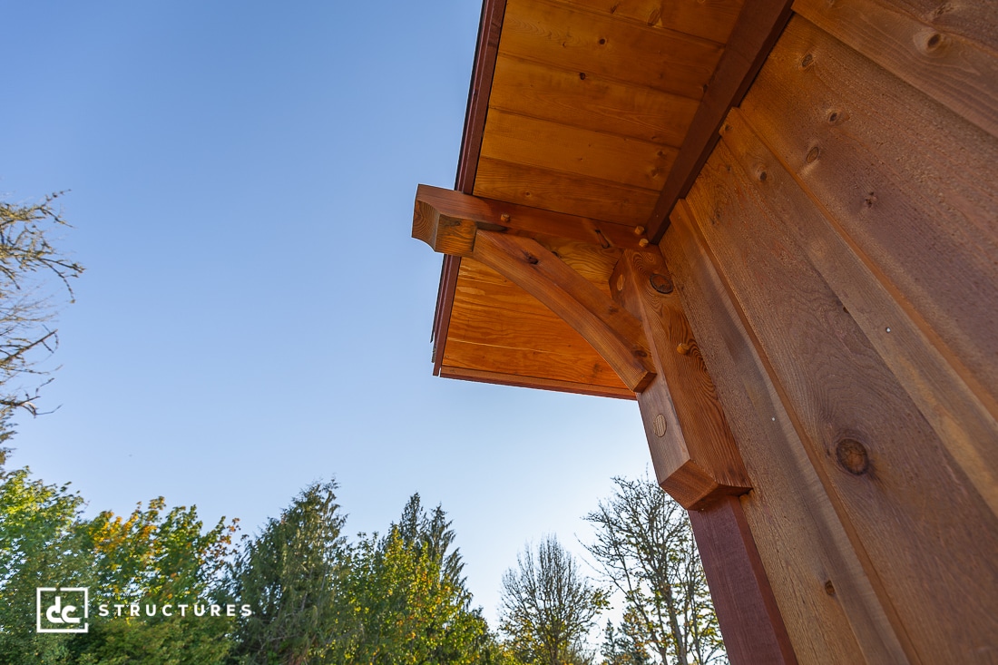 Close-up view of the corner roof and wooden exterior of a building, with clear blue sky and green trees in the background.