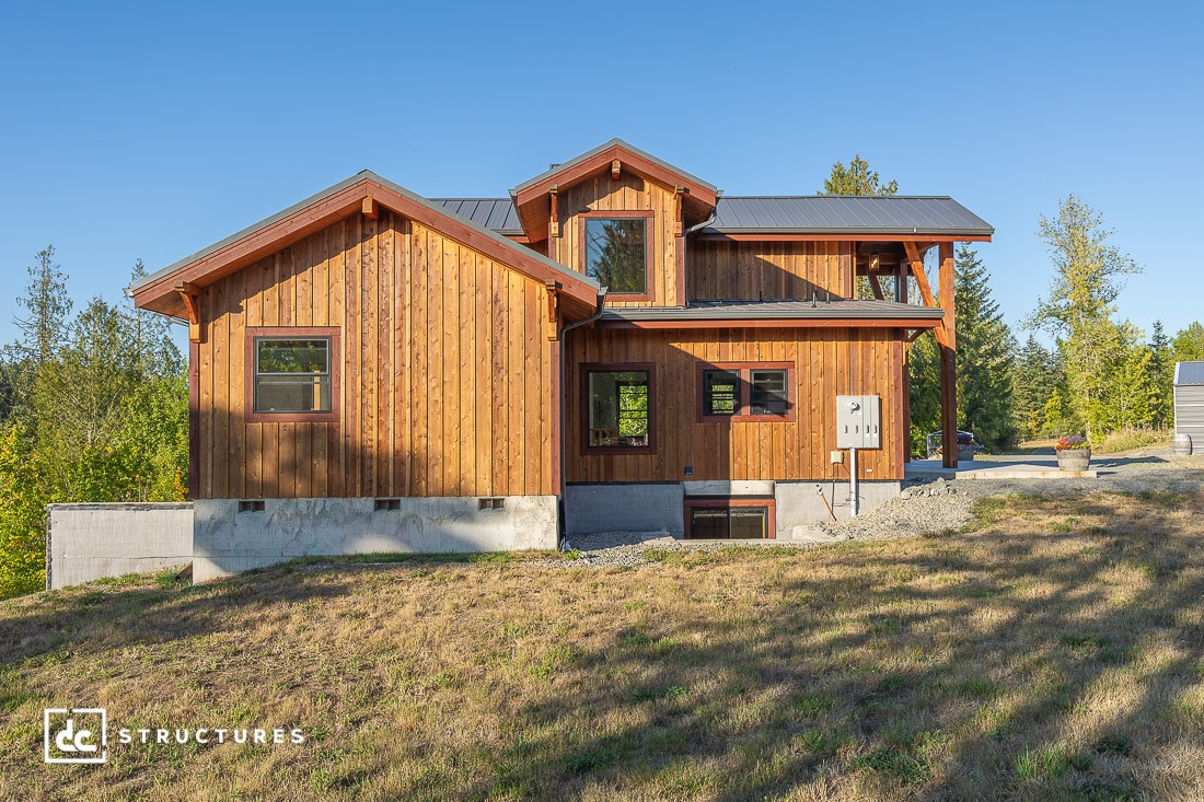 A two-story wooden house with a metal roof sits on a grassy, sloped plot surrounded by trees. The home has large windows and a visible concrete foundation. The sky is clear and sunny.
