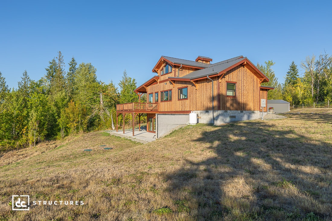 A modern wooden house with large windows and a wraparound deck sits on a grassy hill surrounded by trees, under a clear blue sky.