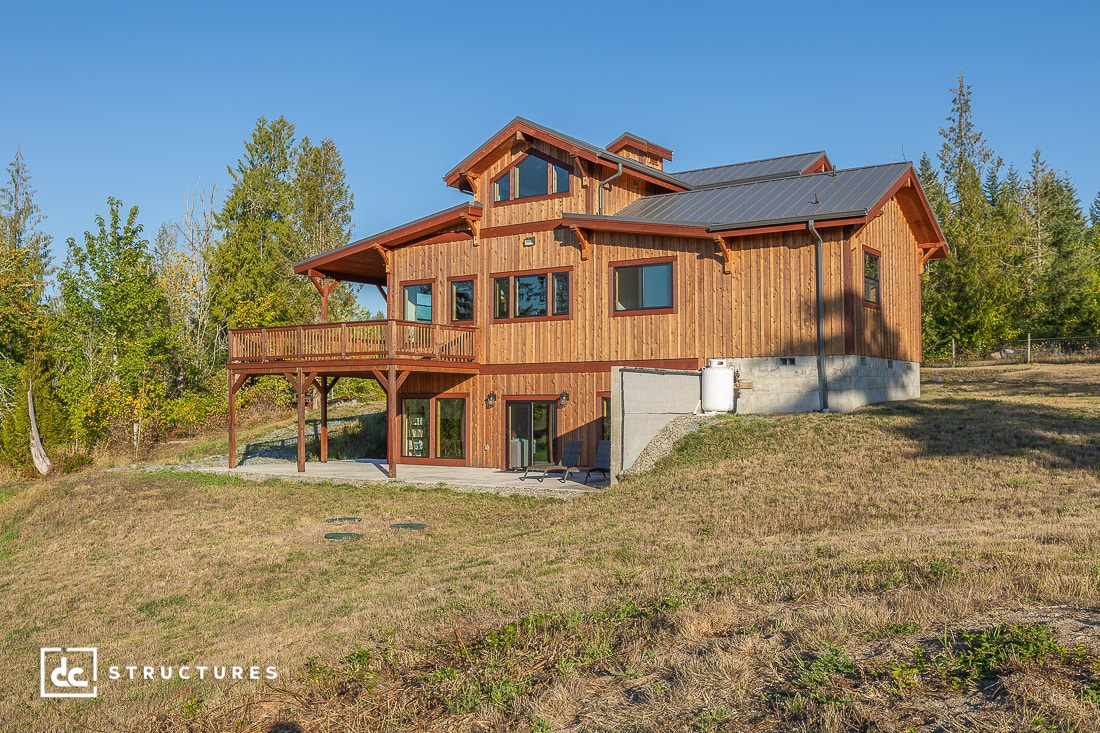 A large rustic wooden house with multiple gabled roofs and a wraparound balcony sits on a sloped grassy plot under a blue sky.