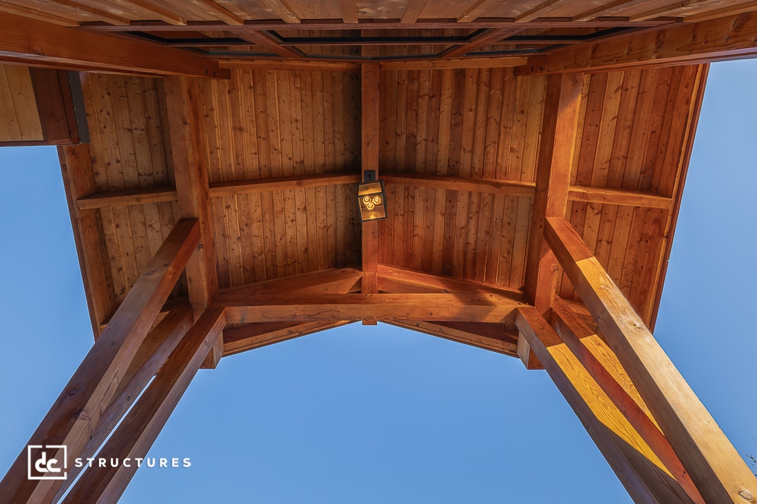 Upward view of a wooden overhang with exposed beams and a light fixture, framed by tall wooden posts against a clear blue sky.