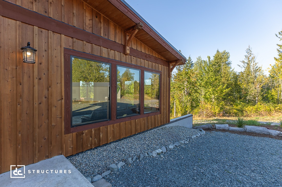 A wooden building with large windows sits on gravel, surrounded by trees and greenery under a clear blue sky.