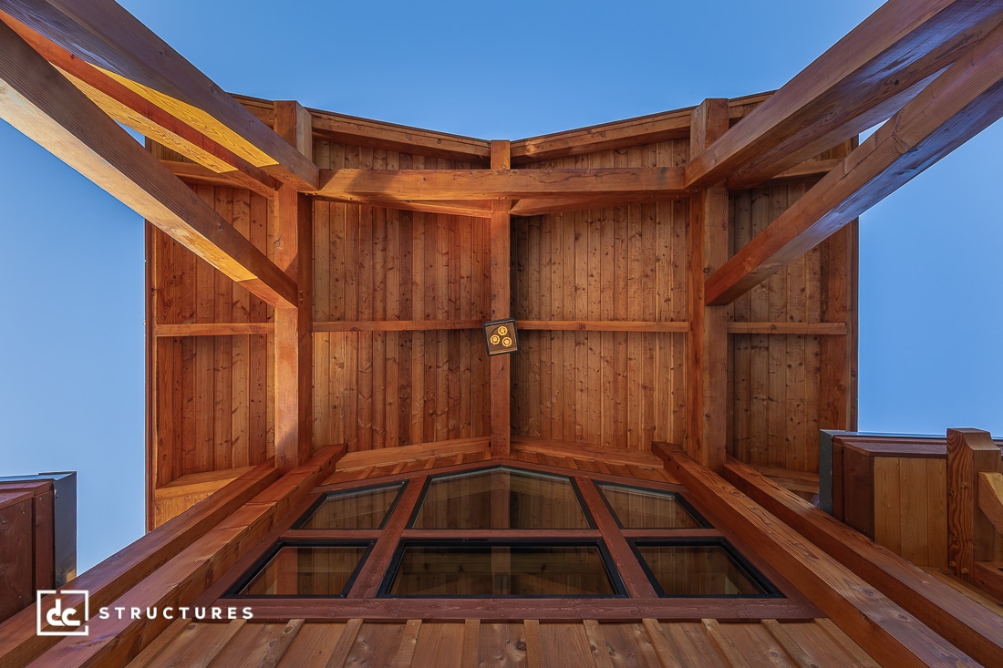View looking up at a wooden pergola or porch roof with exposed beams and panels, set against a clear blue sky. Large windows visible.