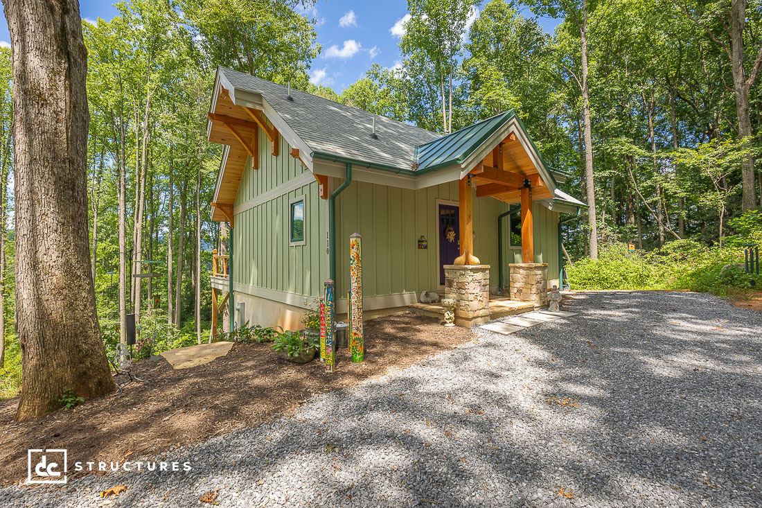 A small green cottage with a gabled roof sits among tall trees, featuring a porch with wooden posts, stone bases, and a gravel driveway.