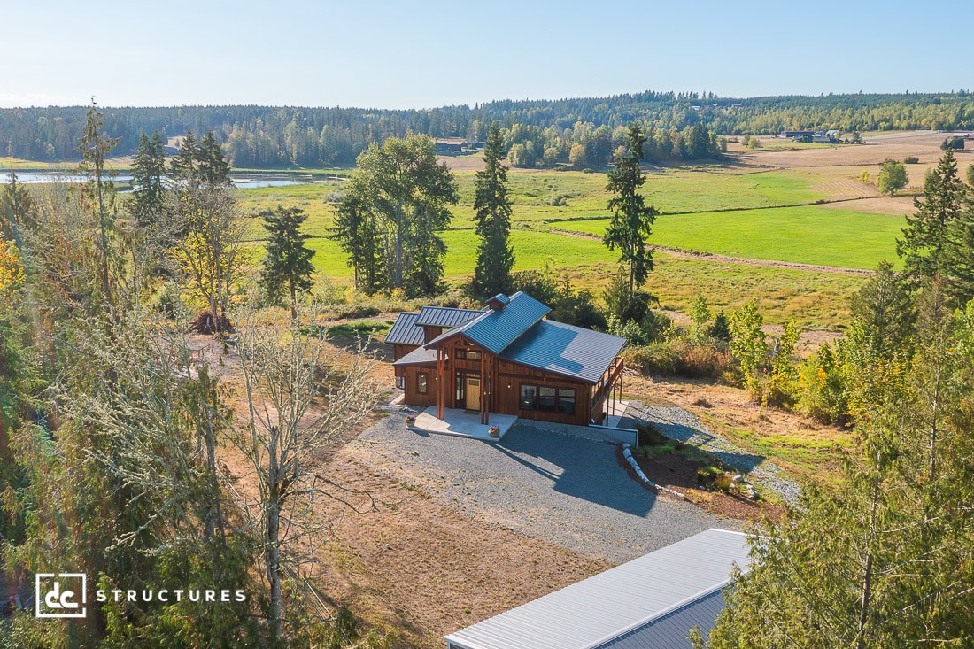 A small wooden cabin with a black roof sits in a rural landscape surrounded by trees, open fields, and distant forest under a clear blue sky.