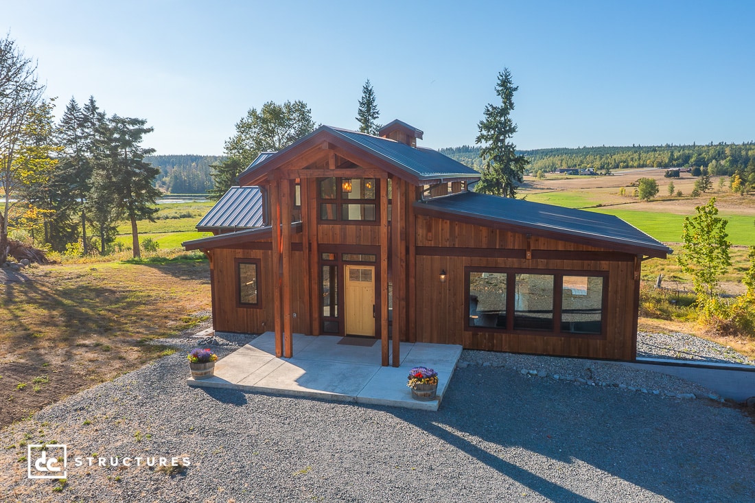 A two-story modern barn-style house with wood siding, large windows, and a covered porch in a rural landscape under a blue sky.