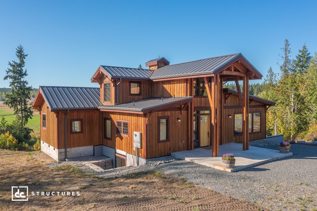 A modern two-story wooden house with a metal roof sits among trees and grass. Flower pots are by the entrance and a gravel driveway in front.