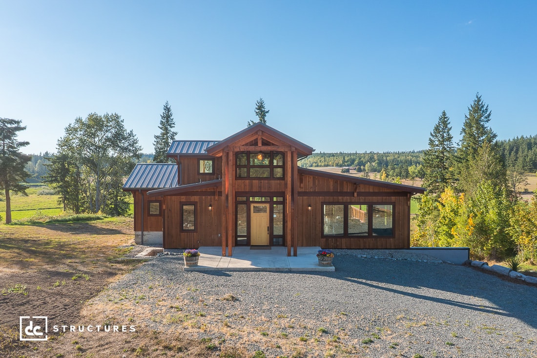 A modern rustic wooden house with large windows and a metal roof, surrounded by trees and fields under a clear blue sky.