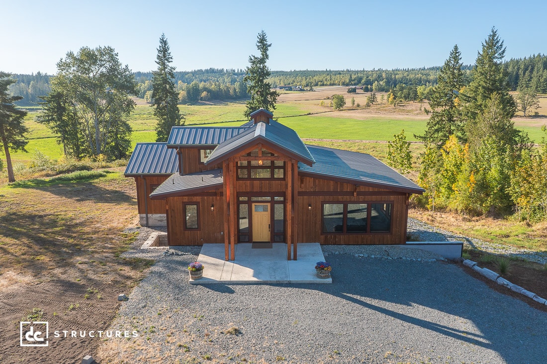 A modern wooden house with large windows and metal roof sits among trees and open fields under a clear blue sky, with a gravel driveway.