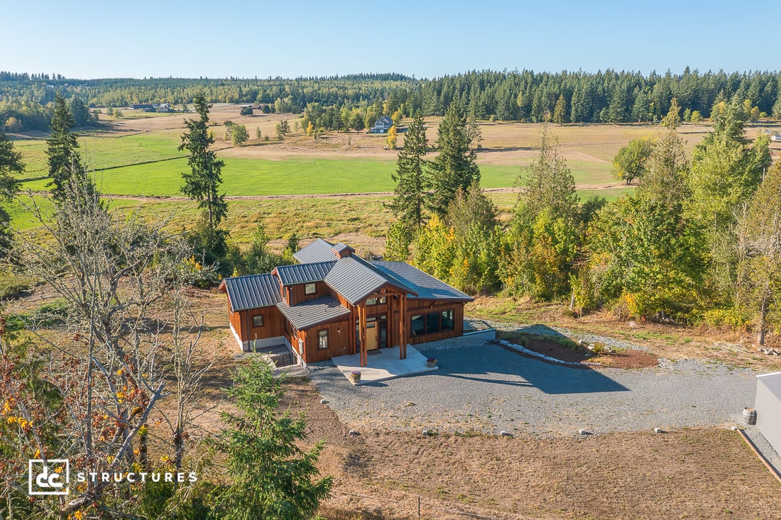 A modern wooden house with a metal roof sits in a clearing surrounded by trees and open fields under a clear blue sky, viewed from above.