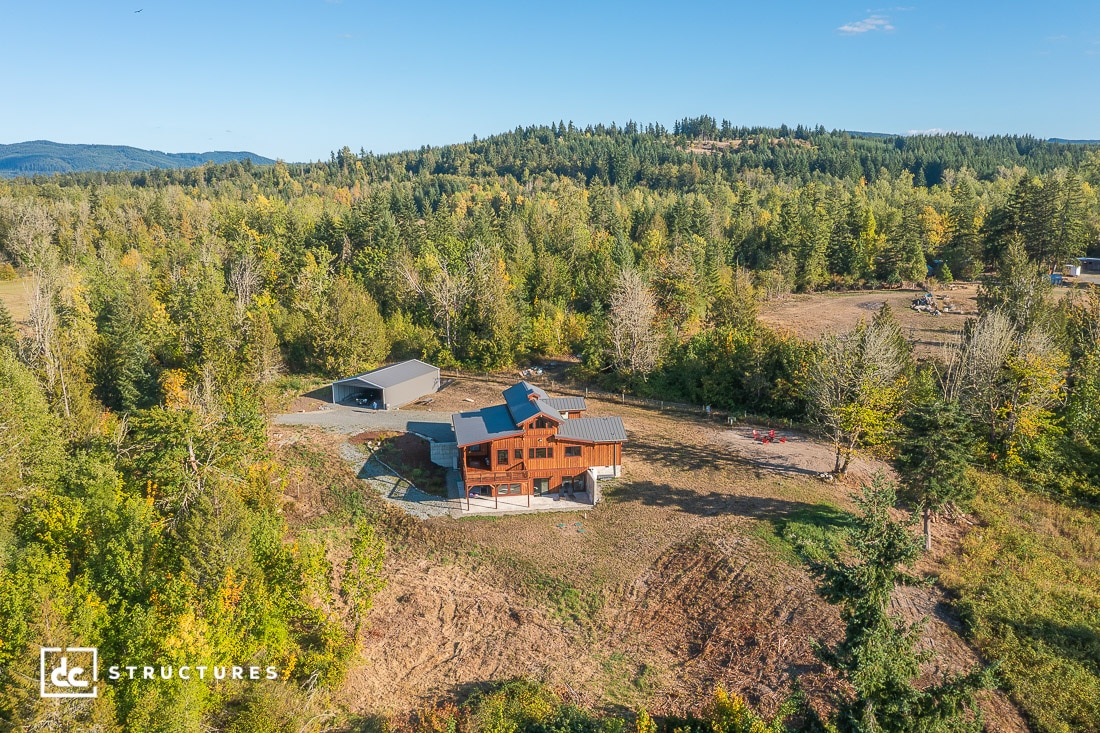 Aerial view of a wooden house with a spacious yard, surrounded by trees and fields, large garage nearby, forested hills behind.