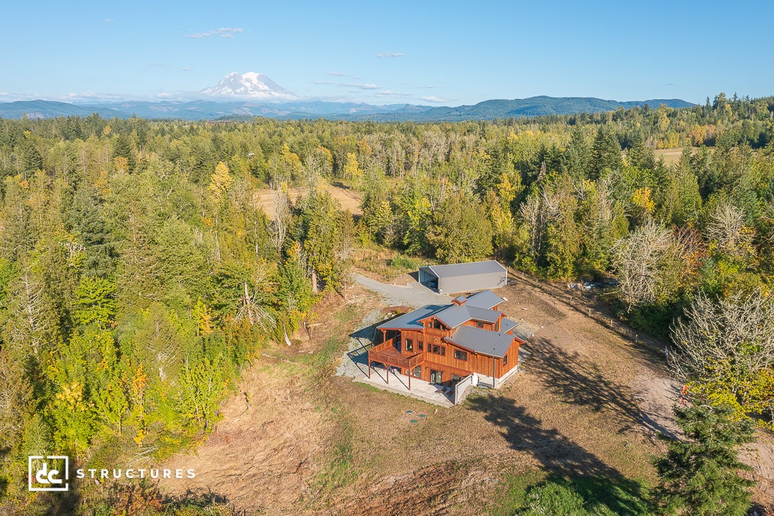 A modern wooden house with outbuildings sits in a clearing surrounded by dense forest, with a snow-capped mountain in the distance under a blue sky.