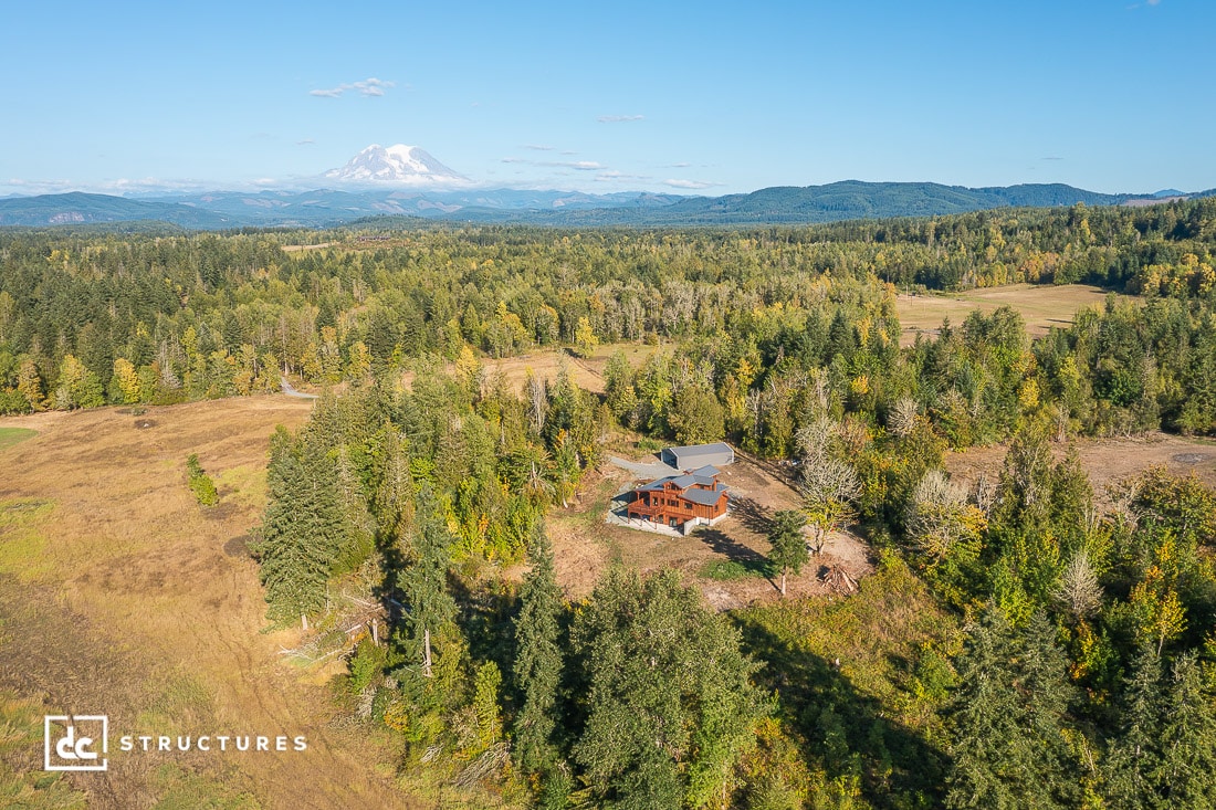 A wooden house surrounded by trees and open fields sits in a rural landscape with a distant snow-capped mountain under a clear blue sky.