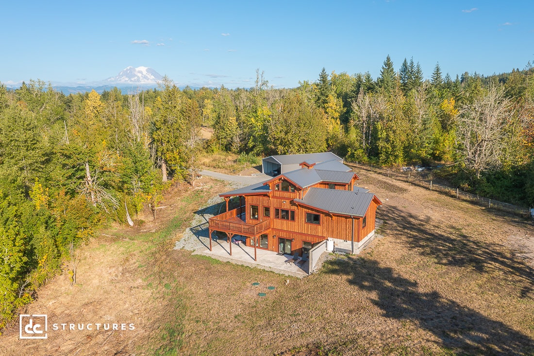 Aerial view of a modern two-story wooden house with gray metal roof, surrounded by trees and open land, mountain in distance.