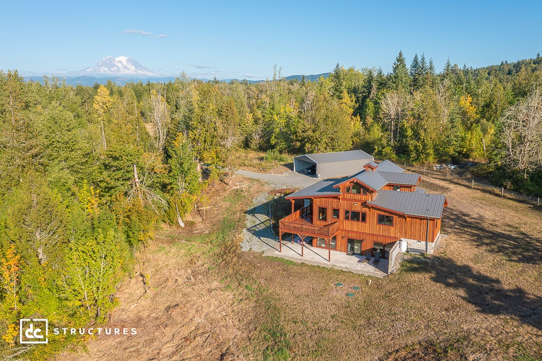 Aerial view of a modern rustic wooden house with covered patio, surrounded by trees, open land, and a distant snow-capped mountain.