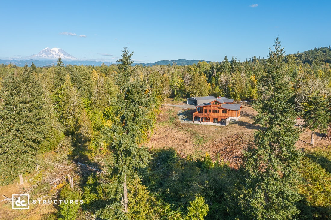 Aerial view of a wooden house among tall evergreens, detached garage nearby, and a snow-capped mountain in the background.