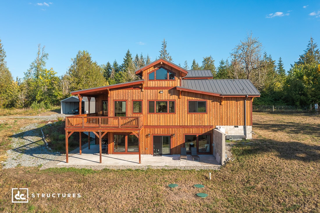 A two-story wooden house with large windows and a covered balcony, surrounded by grass and trees under a clear blue sky.