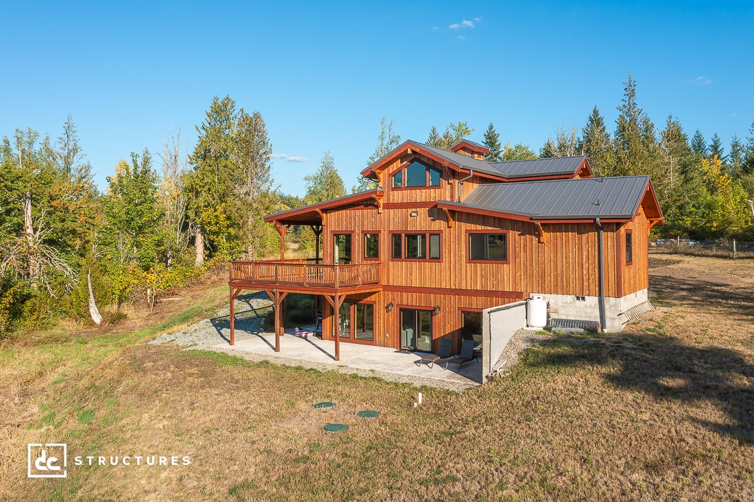 A modern wooden house with large windows and a covered balcony sits on a grassy hillside surrounded by trees under a blue sky.