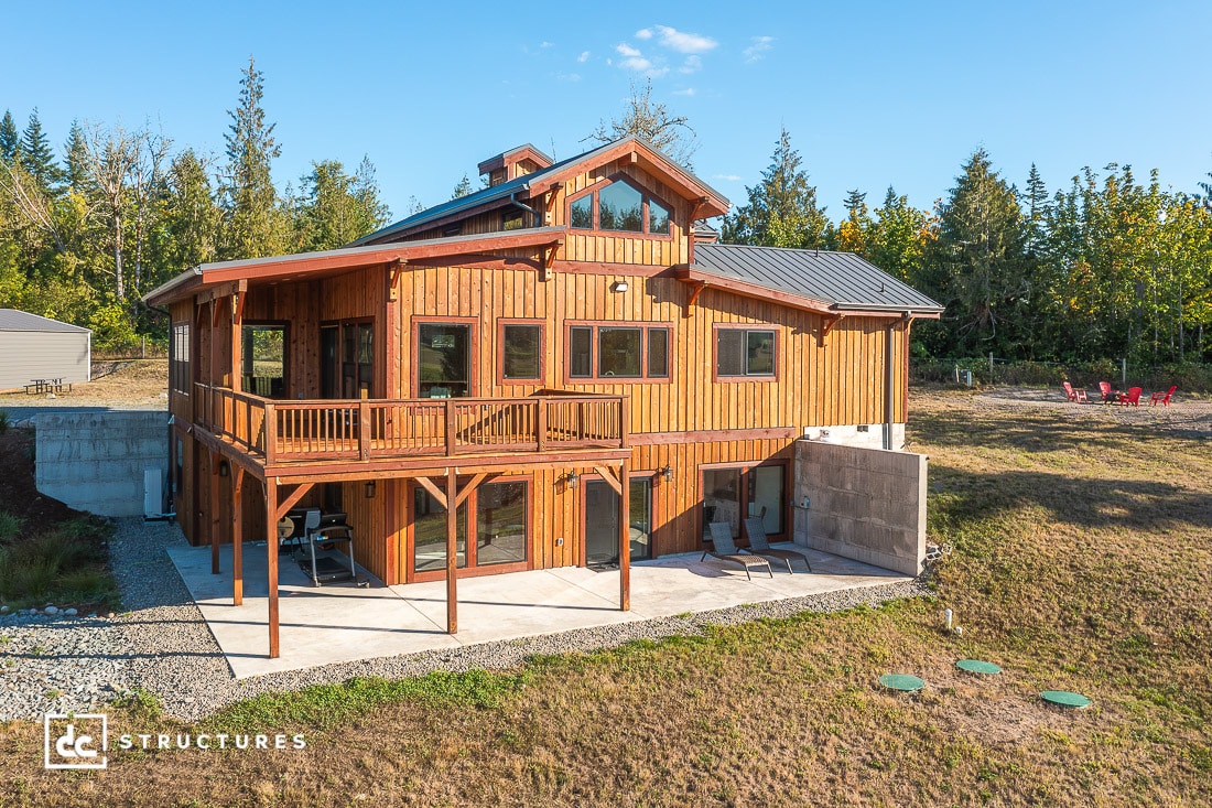 A modern two-story wooden house with large windows, a balcony, and covered patio sits on a grassy lot surrounded by trees under a clear blue sky.