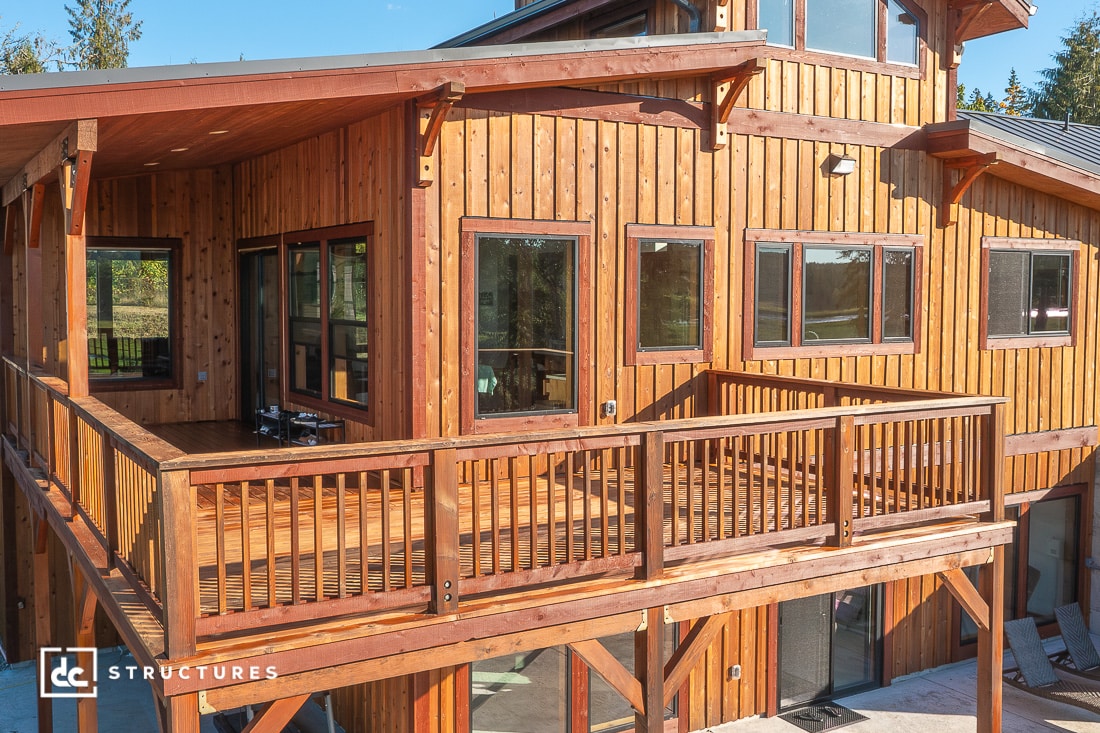 A large wooden house with many windows and a spacious wraparound deck, surrounded by trees and bathed in sunlight.