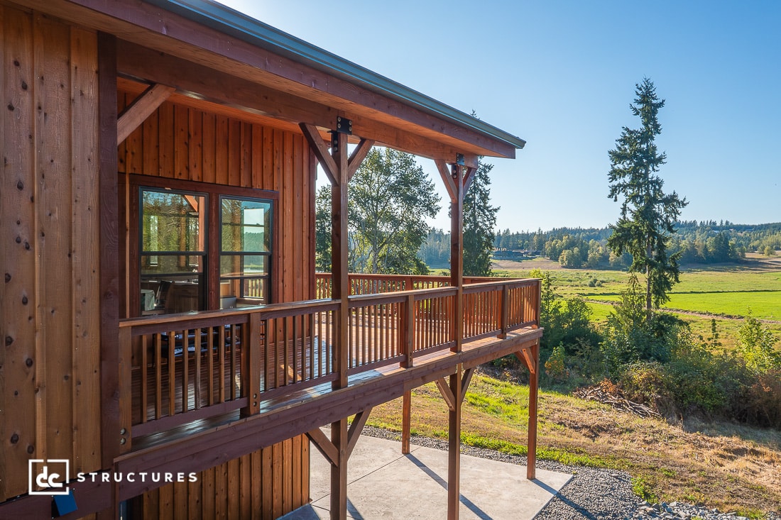 A wooden cabin with a wraparound balcony overlooks green fields and trees under a clear blue sky. The scene is peaceful and sunlit.