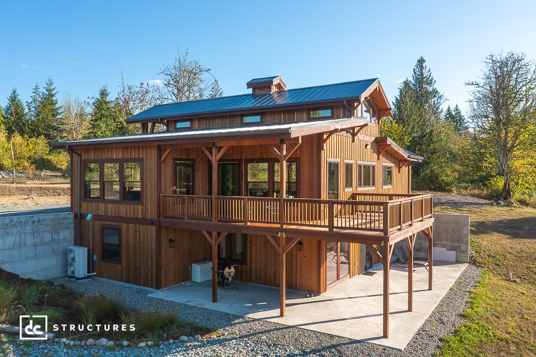 A two-story wooden house with large windows and a wraparound deck in a wooded area, with a metal roof, driveway, and patio.