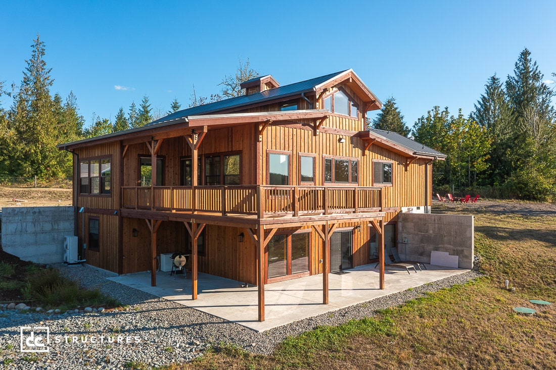 A two-story wooden house with large windows, a covered upper deck, and open lower patio, surrounded by trees and grassy yard.