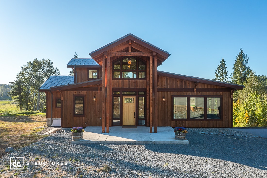 A modern wooden house with large windows and a peaked roof stands on a gravel driveway, surrounded by trees under a blue sky.
