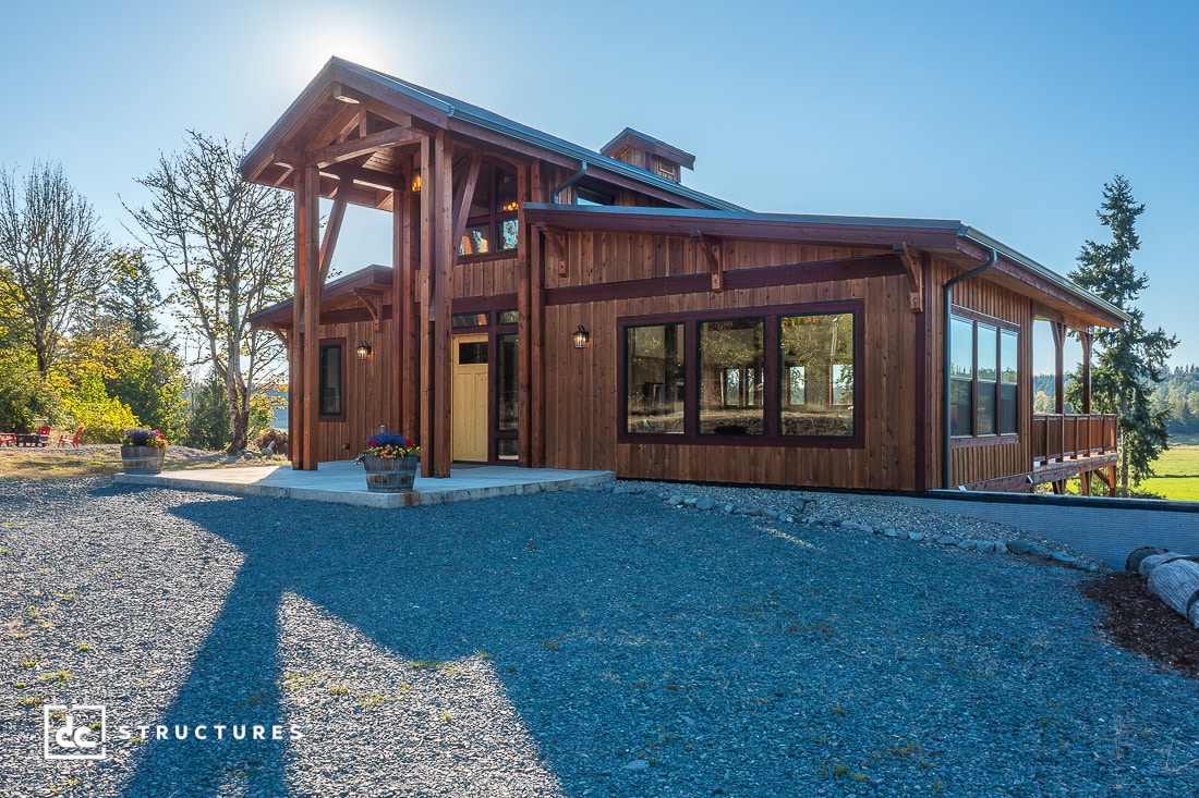 A modern wooden house with large windows and a sloped metal roof sits on a gravel driveway, surrounded by trees and open fields.