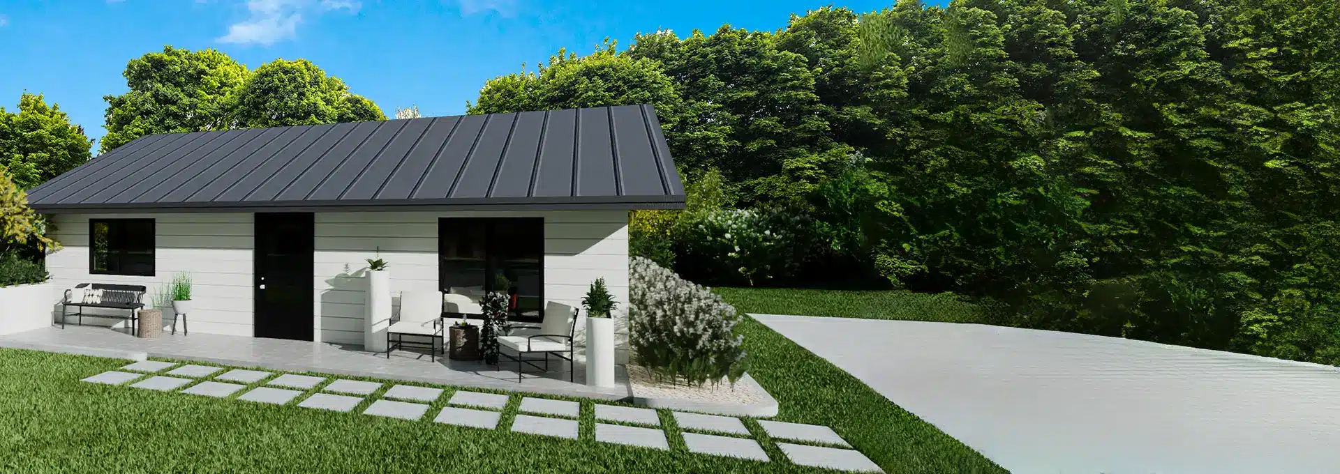 A small modern white house with a dark metal roof sits beside a paved walkway, surrounded by lush grass, plants, and trees.