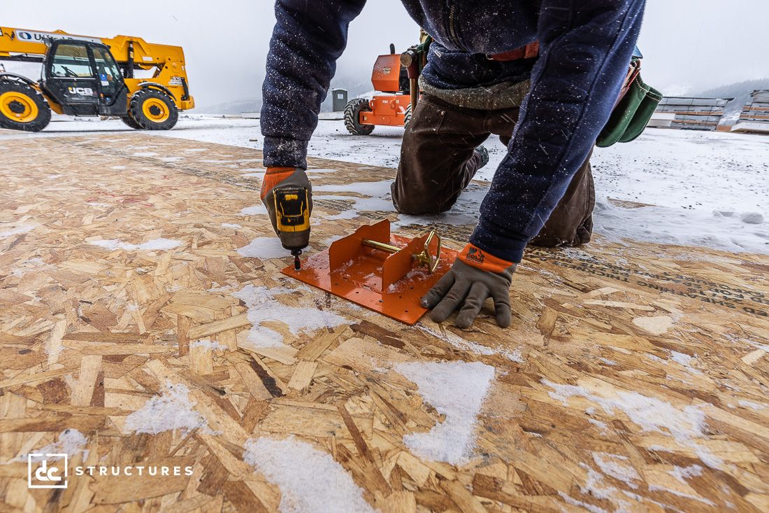 A worker kneels on an OSB plywood floor, using a power drill and metal template outdoors in snowy weather. Construction equipment and snow-covered ground are visible in the background.