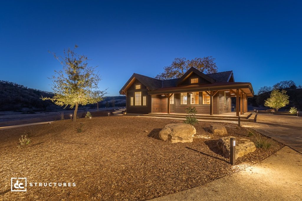 A cozy, single-story wooden house with warm lights glowing inside sits on a landscaped plot with rocks and young trees at dusk. The sky is deep blue.