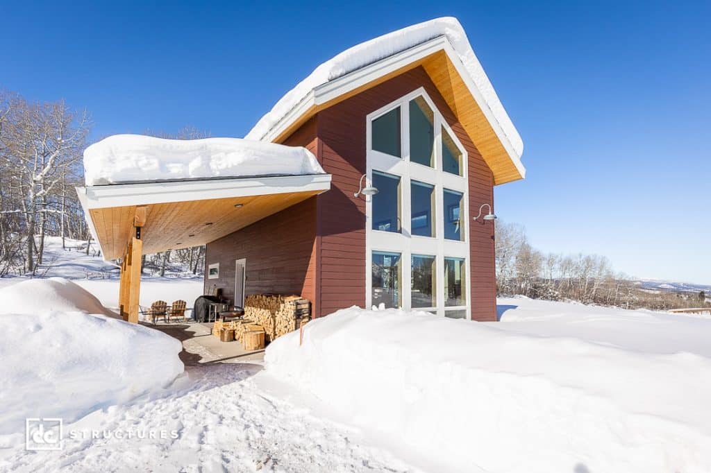 A modern cabin with large windows and a sloped roof is surrounded by deep snow. Stacked firewood sits under its porch.